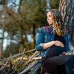Schwangere Frau sitzt im Wald auf Holzstämmen, trägt schwarzes Kleid und Jeansjacke, wirkt entspannt